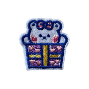 May include: White embroidered patch of a bear wearing a pink bow in a blue and purple gift box.