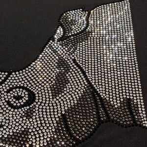 Op de afbeelding: Een zwarte stoffen achtergrond met een ontwerp gemaakt van zilveren en zwarte strass steentjes. Het ontwerp is een gestileerde afbeelding van een gezicht.