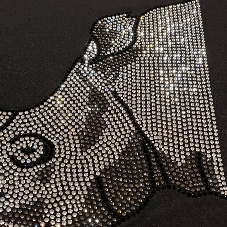 Op de afbeelding: Een zwarte stoffen achtergrond met een ontwerp gemaakt van zilveren en zwarte strass steentjes. Het ontwerp is een gestileerde afbeelding van een gezicht.