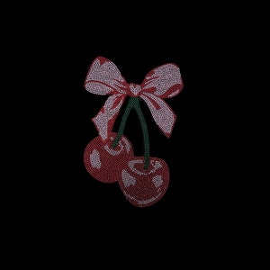 Puede incluir: Dos cerezas rojas con tallos verdes y un lazo rosa. Las cerezas y el lazo están hechos de diamantes de imitación.