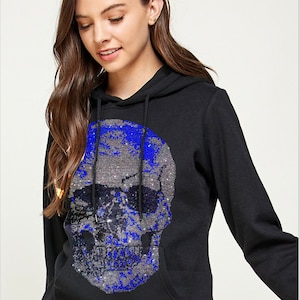 Könnte beinhalten: Schwarzer Hoodie mit einem großen Totenkopf-Design aus Strasssteinen mit blauen Akzenten. Der Totenkopf trägt eine Sonnenbrille.