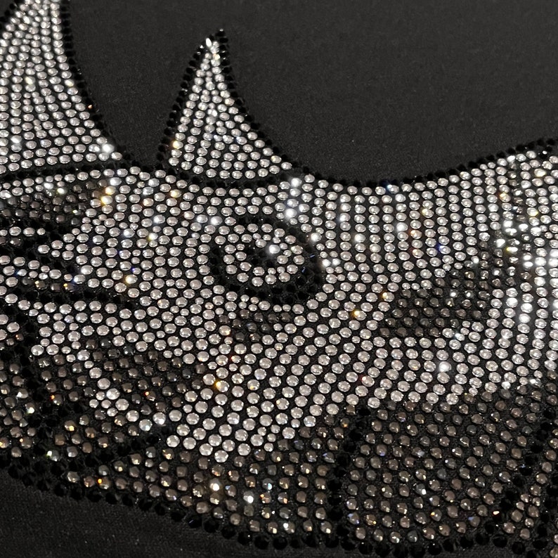 Op de afbeelding: Een zwarte stoffen achtergrond met een ontwerp van strasssteentjes in de vorm van een neushoorn. De strasssteentjes zijn zilver en zwart.