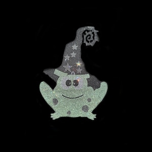 Può includere: Una rana di Halloween ricoperta di strass con un cappello da strega. La rana è verde chiaro con macchie verde scuro e grandi occhi rotondi. Il cappello da strega è nero con accenti di stelle e un disegno a ragnatela. L'immagine è su sfondo nero.