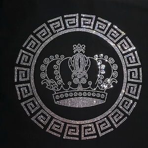 Può includere: Una maglietta nera con un design di corona in strass argentato all'interno di un cerchio di motivi a chiave greca.