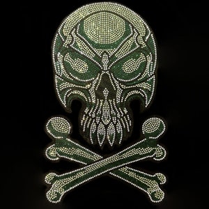 May include: A green and white skull and crossbones design made of rhinestones. The skull has a detailed design with a pointed chin and a large, open mouth. The crossbones are simple and symmetrical.