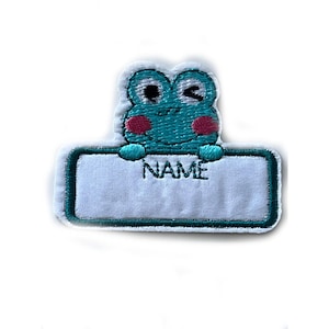 Puede incluir: Una rana verde con mejillas rosadas sostiene una etiqueta de nombre blanca con la palabra "NAME" bordada.