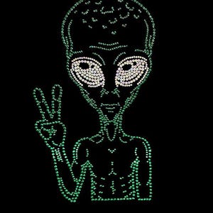 May include: A green alien design on a black background. The alien is outlined with green rhinestones, with white rhinestones for the eyes. The alien is giving a peace sign with its hand.
