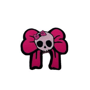 May include: Pink fabric patch with a skull wearing a pink bow. The skull has black eyes and a white outline.