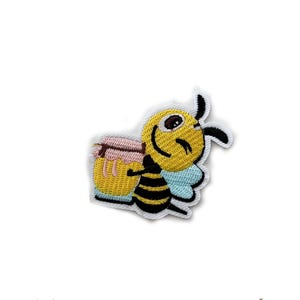 Puede incluir: Parche bordado con una abeja de dibujos animados sonriente, sosteniendo un tarro de miel. La abeja es amarilla y negra con alas azul claro. El tarro de miel es amarillo con una tapa rosa. El parche tiene un borde blanco.