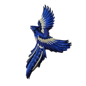Könnte beinhalten: Blaue und goldene gestickte Vogel-Applikations-Patch. Der Vogel ist im Flug mit weit ausgebreiteten Flügeln. Der Patch ist perfekt, um Kleidung, Taschen oder anderen Bastelarbeiten einen Hauch von Verspieltheit zu verleihen.