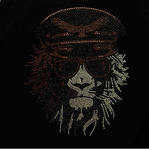 Peut inclure: Un design de tête de lion en strass noir et blanc avec un chapeau et des lunettes de soleil en strass marron. La crinière du lion est faite de strass.