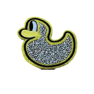Puede incluir: Un parche de fieltro amarillo con un contorno negro en forma de pato de goma. El pato está cubierto de purpurina plateada.