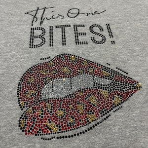May include: Grey t-shirt with the text "This One Bites!" in black rhinestones. A large red, black, and gold rhinestone lip design is below the text.