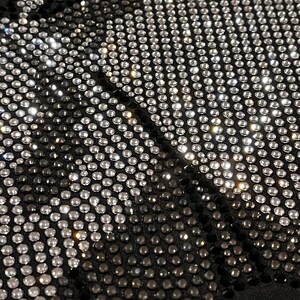 Op de afbeelding: Zwarte stof bedekt met een patroon van zilveren en zwarte strass steentjes. De strass steentjes zijn in rijen gerangschikt en cre&euml;ren een geometrisch ontwerp.