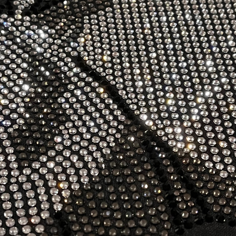 Op de afbeelding: Zwarte stof bedekt met een patroon van zilveren en zwarte strass steentjes. De strass steentjes zijn in rijen gerangschikt en cre&euml;ren een geometrisch ontwerp.