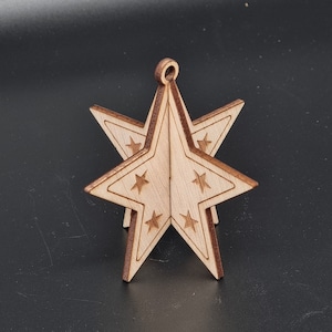 Puede incluir: Un adorno de estrella de madera con un diseño 3D. La estrella es de color marrón claro y tiene un pequeño lazo en la parte superior para colgar. La estrella está decorada con estrellas más pequeñas.