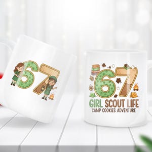May include: Two white ceramic mugs with a design featuring the number 67 in green and brown, with illustrations of Girl Scouts. One mug has the text "GIRL SCOUT LIFE CAMP COOKIES ADVENTURE".
