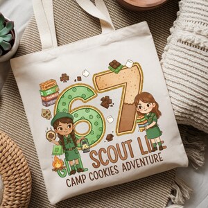 May include: Cream-colored tote bag featuring a colorful illustration with the text "67 Scout Life Camp Cookies Adventure." The design includes cartoon figures of Girl Scouts, cookies, marshmallows, and chocolate bars. The bag has two cream-colored handles.