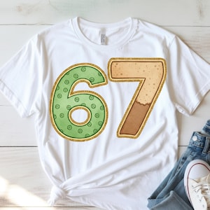 May include: White t-shirt featuring the number 67 in a decorative style. The number 6 is green with a pattern of small circles, while the 7 is tan with a brown base. Both numbers have a gold glitter outline.