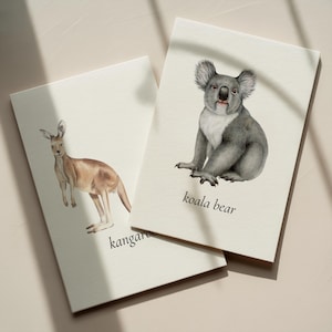 May include: Two cream-colored cards with watercolor illustrations of Australian animals. One card features a kangaroo, the other a koala bear. The koala card has the words "koala bear" printed below the image. The kangaroo card has the word "kangaroo" printed below the image.