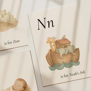 Pode incluir: Ilustração em aquarela de uma impressão do alfabeto infantil com a letra "Nn" e uma representação da Arca de Noé, girafas e elefantes. O texto "is for Noah's Ark" está abaixo da imagem. Outras impressões com "is for Zion" e um bebê dormindo também são visíveis.