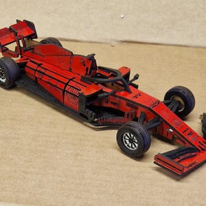Pode incluir: Um modelo de madeira vermelho e preto de um carro de corrida de Fórmula 1. O carro apresenta detalhes em preto, incluindo os pneus, o chassis e os elementos aerodinâmicos. O modelo é uma réplica detalhada de um carro de corrida.