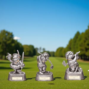 May include: Three silver-colored golf trophies on a green golf course. Each trophy features a whimsical golfer in different poses, including one with a snorkel and fish. Each trophy has a rectangular base with space for engraving.