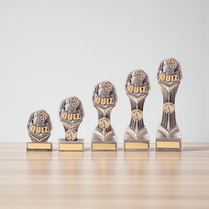 Puede incluir: Un conjunto de cinco trofeos de concurso de diferentes alturas. Cada trofeo presenta una combinación de colores plateado y dorado, con la palabra "QUIZ" en un lugar destacado. Los trofeos tienen un diseño detallado y están colocados sobre una superficie de madera.