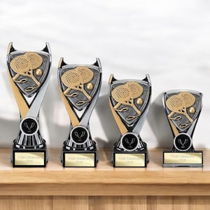 May include: Four silver and gold trophies of varying heights, each featuring a central emblem of two padel rackets and a ball. The base of each trophy includes a rectangular gold plate with the printed text "Your Engraving Here".