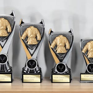 Op de afbeelding: Vier vechtsporttrofeeën met een gouden gi-ontwerp op een zilver-zwarte basis. Elke trofee heeft ruimte voor graveren. De trofeeën worden gepresenteerd op een houten oppervlak.