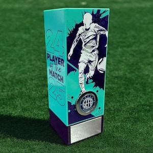 May include: A tall, rectangular trophy with a teal and dark purple colour scheme. It features a graphic of a football player, the text 24 PLAYER OF THE MATCH 25, a circular SEFA logo emblem, and a blank silver rectangular plate at the base.