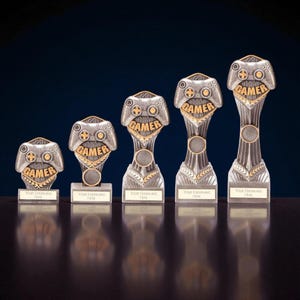 May include: A collection of silver and gold-colored gamer trophies. Each trophy features a controller design with the word "GAMER" and space for engraving. The trophies vary in size and are displayed on a reflective surface.