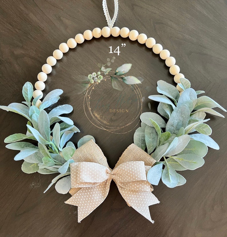 May include: A 14 inch wood bead wreath decorated with light green lamb's ear greenery and a beige burlap bow with white polka dots.