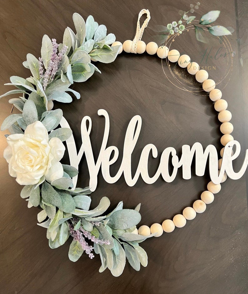 May include: A welcome wreath with a white wooden welcome sign, a white flower, and green leaves. The wreath is made of wooden beads and has a white ribbon for hanging.