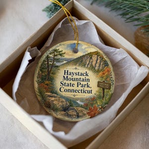 May include: A round ceramic ornament featuring a scenic landscape of Haystack Mountain State Park, Connecticut. The ornament is in a gift box with a gold string for hanging. The design includes a mountain, trees, and the park's name.
