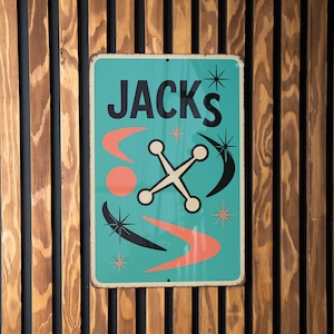 May include: A retro-style teal metal sign with the word "JACKS" in black lettering. The sign features mid-century modern design elements, including a white cross-shaped design, orange accents, and starbursts. The sign is mounted on a wooden slatted wall.