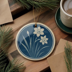 May include: Round ceramic ornament featuring a floral design of white daffodils on a deep blue background. The ornament has a cream-colored border and a gold string for hanging. A decorative item for the home.
