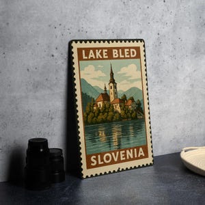 May include: A vintage-style metal sign featuring a scenic view of Lake Bled, Slovenia, resembling a postage stamp. The sign displays a church on an island, surrounded by trees, with the text "LAKE BLED" and "SLOVENIA". The color palette includes shades of brown, blue, and green.