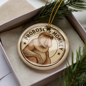 May include: A round, cream-colored Christmas ornament featuring a detailed illustration of a proboscis monkey. The text "PROBOSCIS MONKEY" is printed around the monkey image in a brown circular border. The ornament is in a gift box with a gold string.