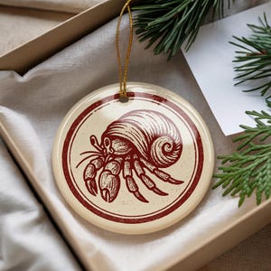May include: A round, cream-colored ceramic ornament featuring a detailed illustration of a hermit crab in a dark red color. The crab is enclosed within a dark red circular border. A gold string is attached for hanging.