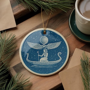 May include: Round ceramic ornament with a blue background featuring a white illustration of an Egyptian figure in a boat, with wings and a sun. The ornament has a gold string for hanging. The border is off-white.