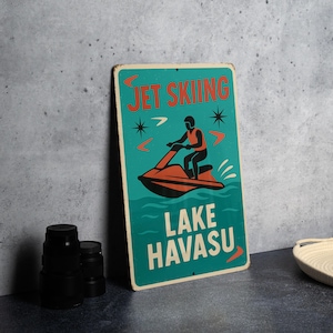 May include: Vintage metal sign with a teal background featuring the words "JET SKIING" and "LAKE HAVASU" in white and orange. An illustration of a person riding a jet ski is in the center.