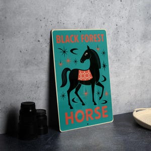 May include: A teal metal sign with a black horse illustration and the words "BLACK FOREST HORSE" in orange. The horse has a red and white patterned blanket. Decorative starbursts and crescent shapes surround the horse.
