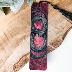 May include: A rectangular bookmark with a dragon design. The dragon is gray and black, with a red and pink background. A black tassel hangs from the top. The bookmark is on a wooden surface.