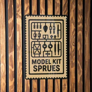 May include: A rectangular sign with a black border and the words "MODEL KIT SPRUES" in black text. The sign features a diagram of model kit parts. The sign is mounted on a wooden wall with vertical brown and black planks.