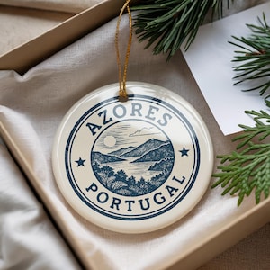 May include: A round, cream-colored ceramic ornament with a blue and white design. The ornament features the text "AZORES PORTUGAL" in a circular border, with a landscape illustration of mountains and water in the center. A gold string is attached for hanging.