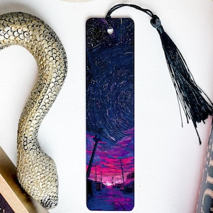 May include: A rectangular bookmark with a night sky design, featuring a swirling galaxy over a street scene with pink and purple hues. It has a black tassel and is next to a book and a decorative snake.
