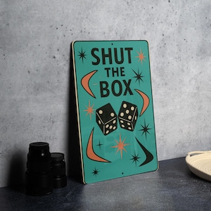 May include: A teal metal sign with the words "SHUT THE BOX" in black, along with dice and starburst designs. The sign has a retro, mid-century modern aesthetic. The dice are black with white dots.