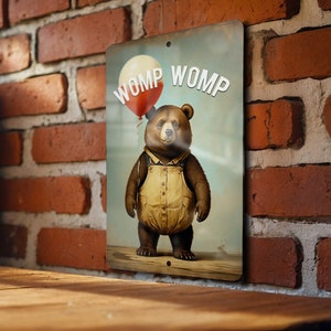 May include: A metal sign with a brown bear wearing overalls and holding a red and white balloon. The sign says "WOMP WOMP" in white letters.