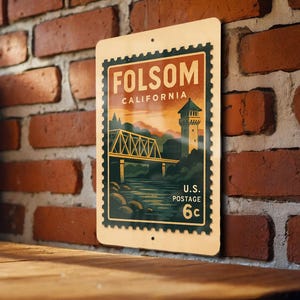 May include: A vintage-style metal sign featuring a scenic illustration of Folsom, California, with a bridge, tower, and river. The sign has a postage stamp design with the text "Folsom California" and "U.S. Postage 6c."